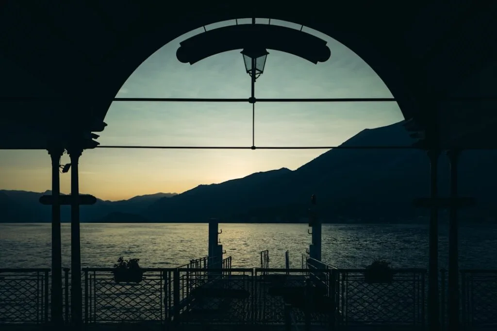 Arcade sombre encadrant un quai et montagnes au coucher sur le Lac de Côme