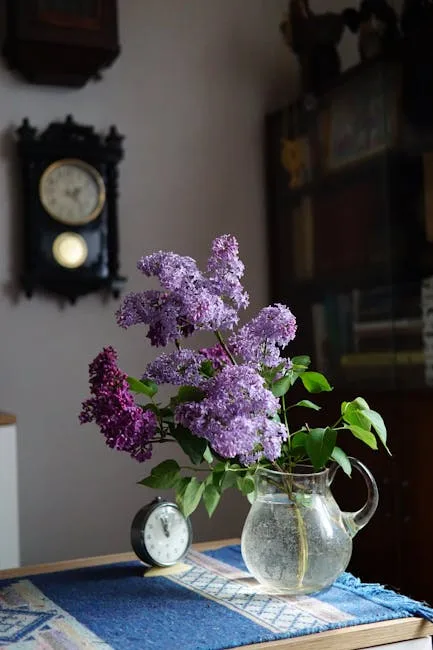Glaskrug mit Fliederblüten auf Tisch, antike Wanduhr im Hintergrund